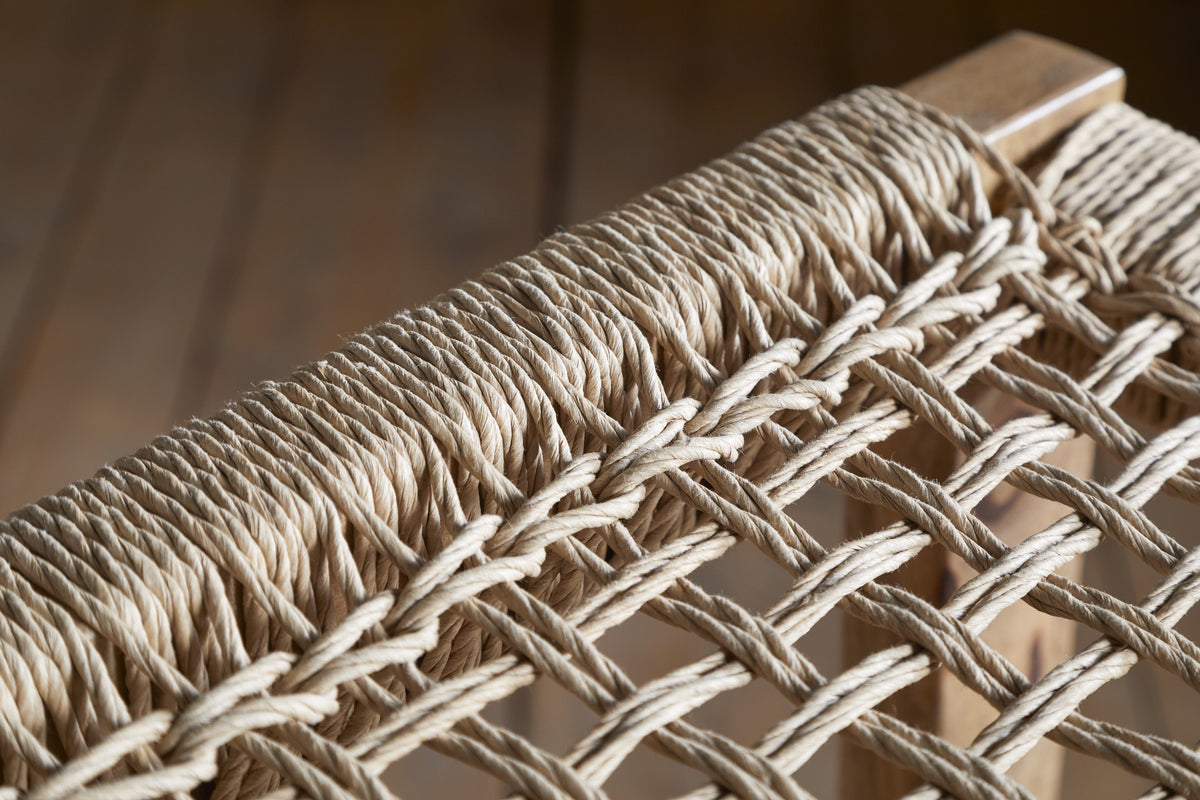 Close-up of the nkuku Aamani Mango Wood & Cord Counter Stool - Natural, featuring a tightly woven natural-colored rope seat on a smooth mango wood frame. Blurred wooden flooring appears in the background.