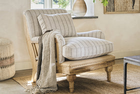 The nkuku Abe Deconstructed Linen Armchair in Natural Stripe features a mango wood frame and comes with a matching pillow and throw, creating a cozy spot on a woven rug. A window, decorative vase, and textured wall art add warmth to the inviting room.