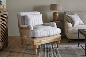 A cozy living room features the nkuku Abe Deconstructed Linen Armchair in natural stripe, boasting a sturdy mango wood frame and light gray cushions. A small round table with a brown lamp, beige sofa, and fringed textured rug complete the look.