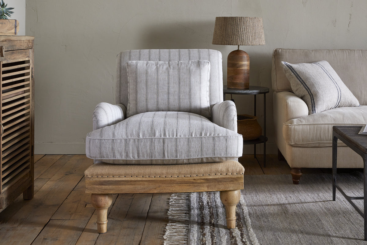 A cozy living room featuring the nkuku Abe Deconstructed Linen Armchair in Stripe Natural with mango wood legs, a matching ottoman, beige sofa with striped cushion, wooden lamp, textured rug, and wood floors for a warm, inviting vibe.