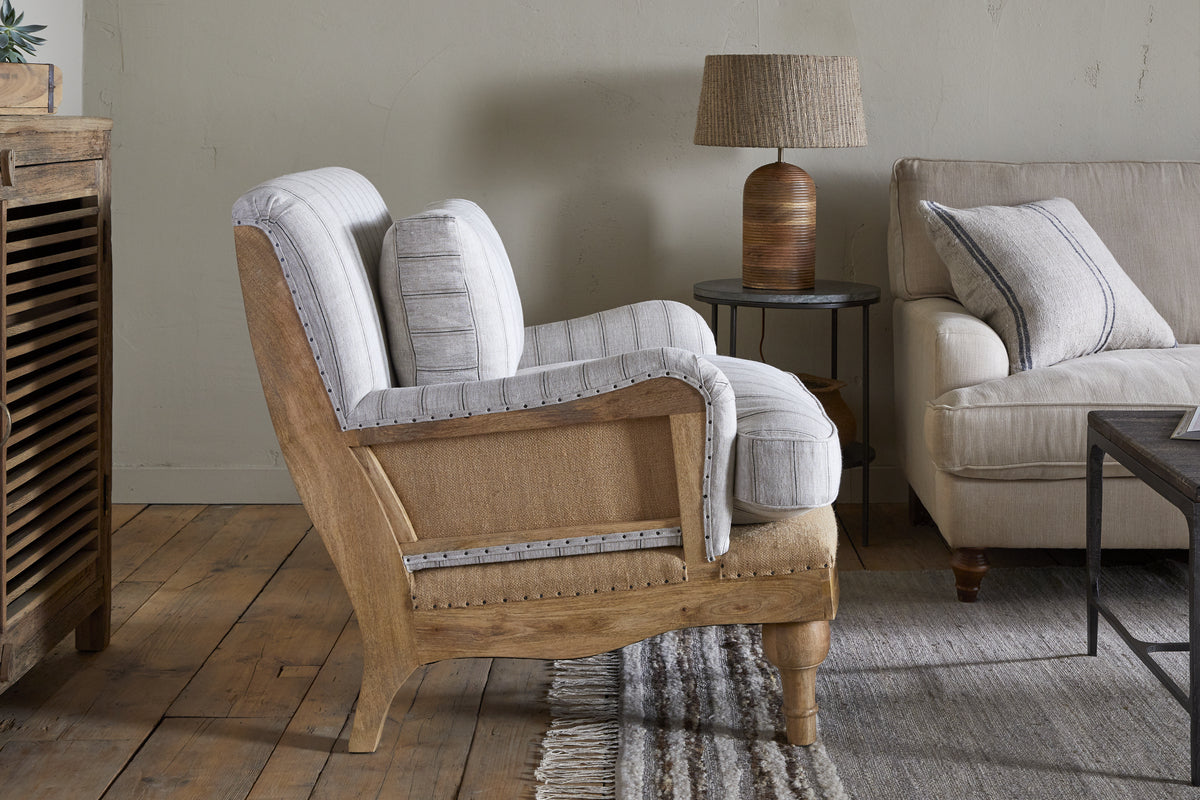 The nkuku Abe Deconstructed Linen Armchair in natural stripe, featuring a mango wood frame and light-gray cushions, is placed on a fringed rug by a round table with a woven lamp, near a beige sofa with a striped pillow.