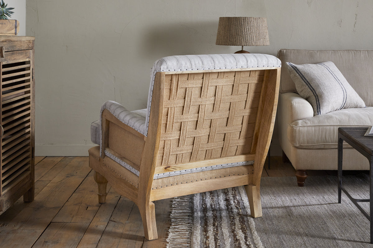 The Abe Deconstructed Linen Armchair - Stripe - Natural by nkuku, featuring a mango wood frame and woven back, faces away on a wooden floor beside a beige sofa, striped rug, textured lamp side table, and wooden cabinet in this cozy living room.