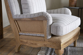 The Abe Deconstructed Linen Armchair - Stripe - Natural by nkuku features a rustic mango wood frame, striped linen upholstery with metal stud accents, and is styled with light beige and gray cushions.