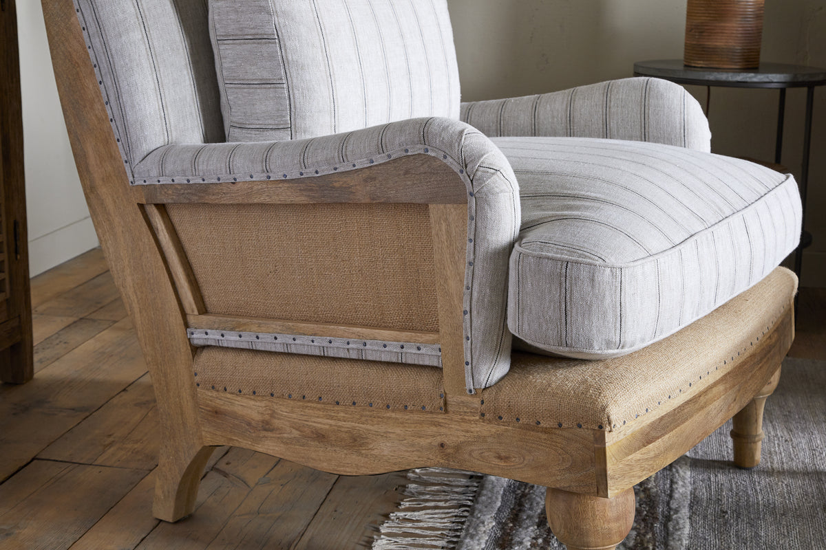 The Abe Deconstructed Linen Armchair - Stripe - Natural by nkuku features a rustic mango wood frame, striped linen upholstery with metal stud accents, and is styled with light beige and gray cushions.