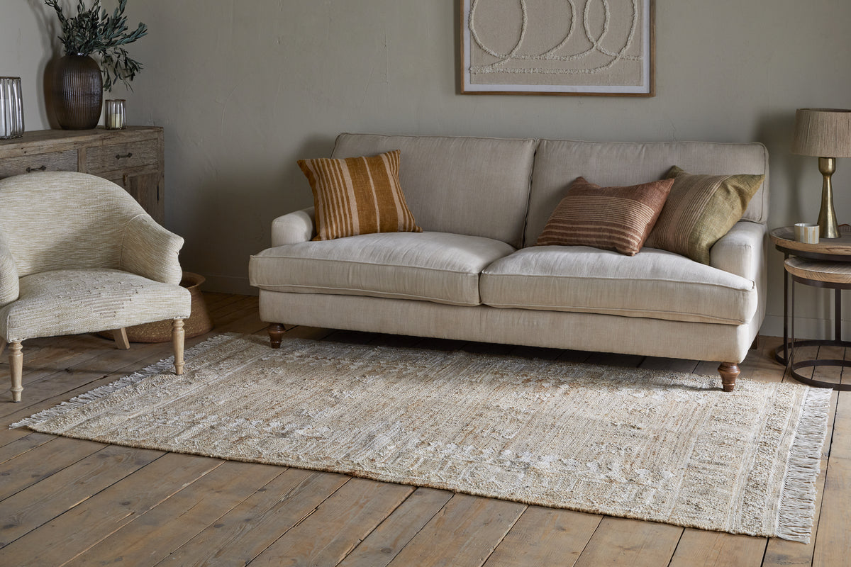 A cozy living room features a beige sofa with brown and green cushions, a nkuku Aqil Jute & Cotton Rug in natural tones on wooden floors, a light armchair, wooden side table, rustic cabinet with vases and greenery, and abstract art on neutral walls.