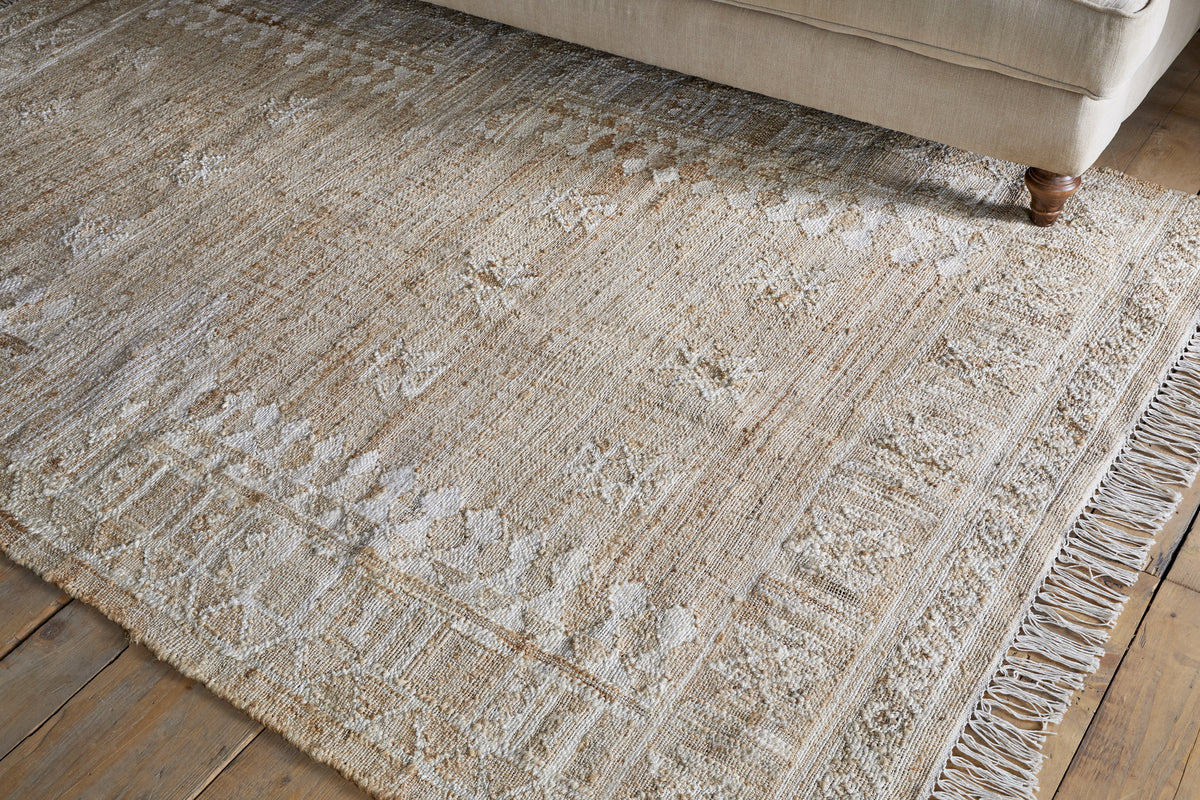 The Aqil Jute & Cotton Rug - Natural by nkuku, featuring traditional Indian geometric and floral patterns in light cream tones, sits on a wooden floor with fringed tassels and a light upholstered sofa partially visible.