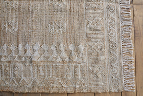 A close-up of the nkuku Aqil Jute & Cotton Rug - Natural reveals woven beige and white geometric patterns, a decorative border with fringed tassels, and highlights its rustic Indian design atop a wooden floor.