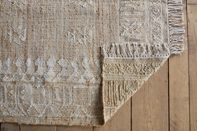 The nkuku Aqil Jute & Cotton Rug - Natural, in beige with white geometric patterns and knotted fringe, lies on a wooden floor. One corner is folded over, showing its thick weave and traditional Indian craftsmanship.