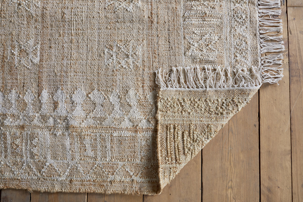The nkuku Aqil Jute & Cotton Rug - Natural, in beige with white geometric patterns and knotted fringe, lies on a wooden floor. One corner is folded over, showing its thick weave and traditional Indian craftsmanship.