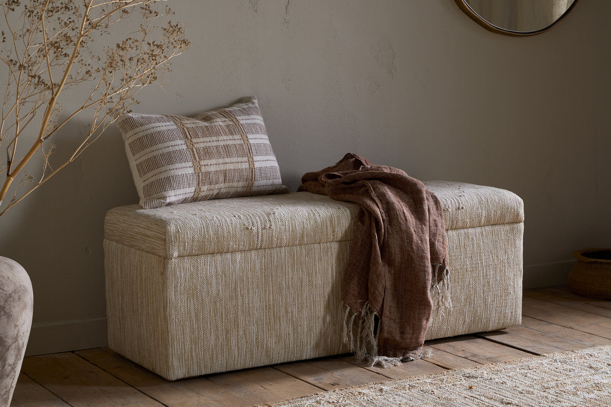 The Aranya Cotton & Jute Upholstered Storage Bench by nkuku sits against a light wall on a wooden floor, topped with a striped beige-and-white pillow and brown fringed throw. A round mirror, dried branches, and woven basket complete the cozy natural decor.