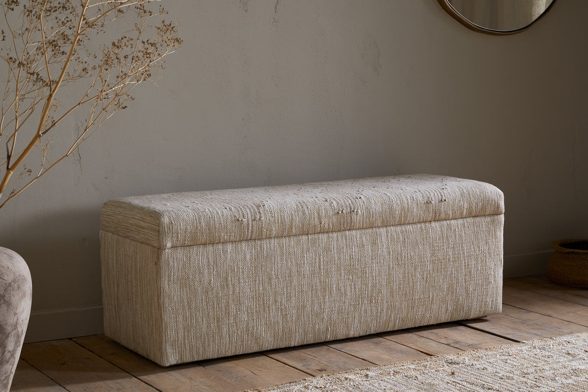 The Aranya Cotton & Jute Upholstered Storage Bench by nkuku sits on a wooden floor against a light gray wall, with dried plant stems in a vase on the left and a round mirror plus woven basket partly visible on the right.