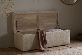 The Aranya Cotton & Jute Upholstered Storage Bench by nkuku in natural sits on a wooden floor, its lid open to show a striped pillow and brown textured blanket. Neutral decor, dried flowers, and a round mirror complete the scene.