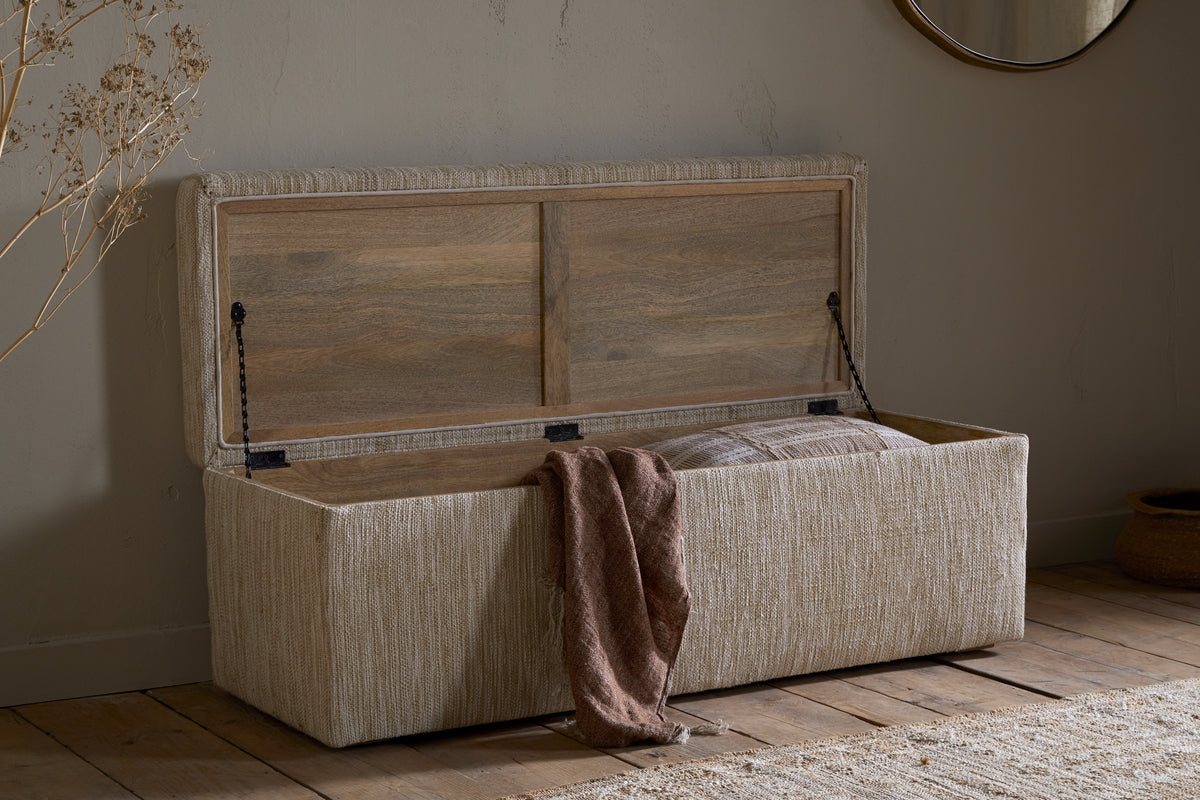The Aranya Cotton & Jute Upholstered Storage Bench by nkuku in natural sits on a wooden floor, its lid open to show a striped pillow and brown textured blanket. Neutral decor, dried flowers, and a round mirror complete the scene.