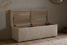 The nkuku Aranya Cotton & Jute Upholstered Storage Bench in Natural sits open against a neutral wall on a wooden floor, complemented by a round mirror, woven rug, and dried plant decor.