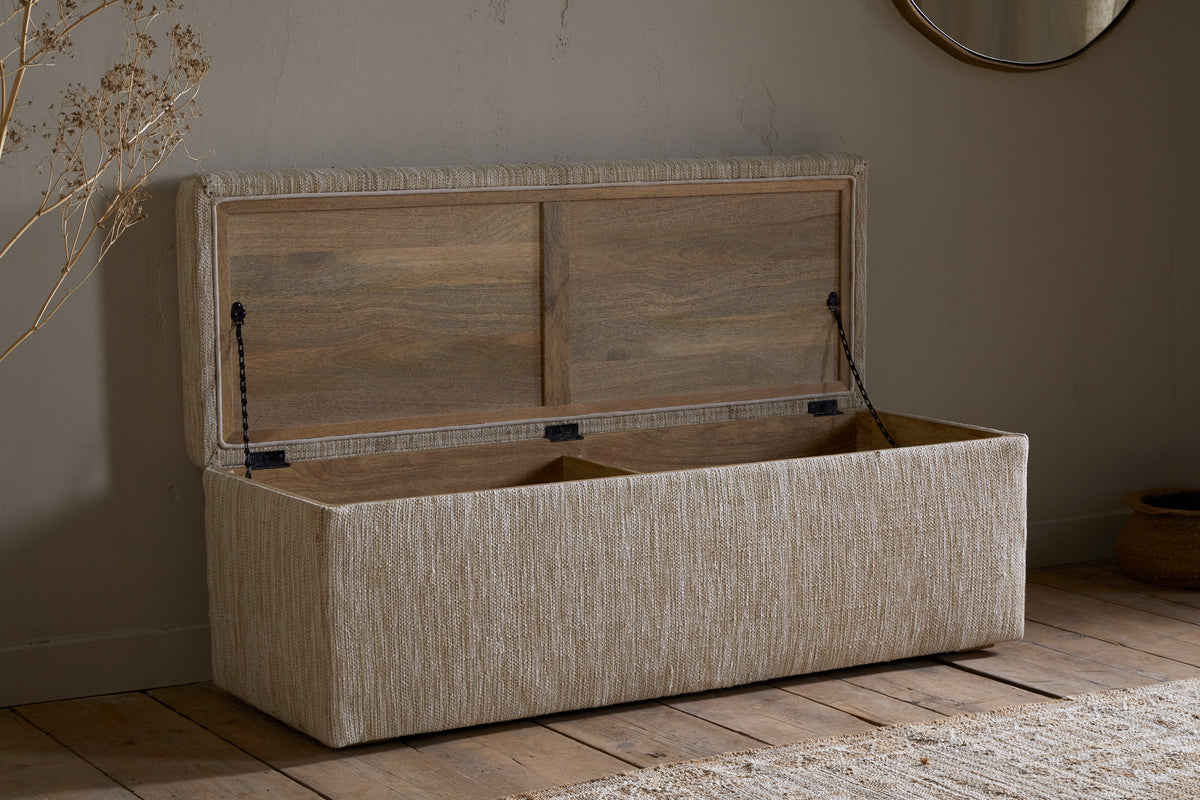 The nkuku Aranya Cotton & Jute Upholstered Storage Bench in Natural sits open against a neutral wall on a wooden floor, complemented by a round mirror, woven rug, and dried plant decor.