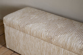 The Aranya Cotton & Jute Upholstered Storage Bench by nkuku features a rectangular design with beige, textured woven fabric and subtle stripes. It sits on wooden floorboards against a plain, light-colored wall.