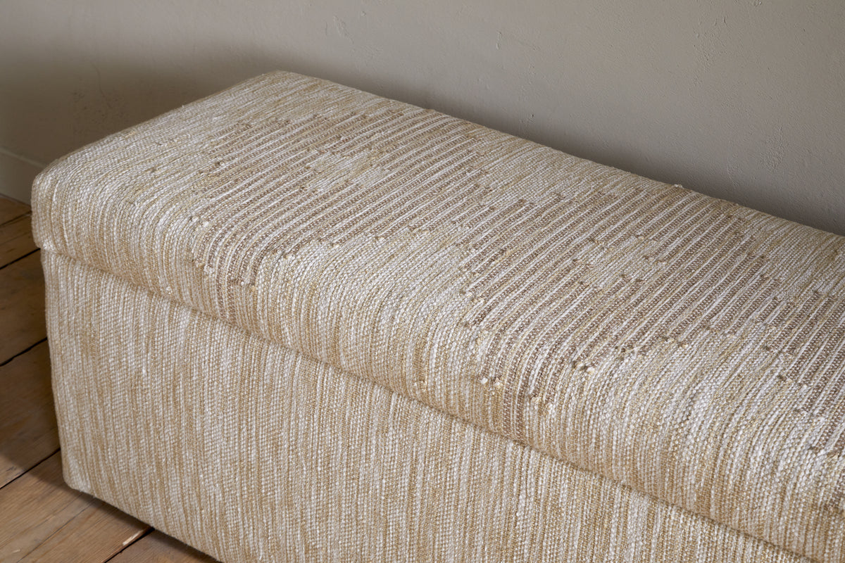 The Aranya Cotton & Jute Upholstered Storage Bench by nkuku features a rectangular design with beige, textured woven fabric and subtle stripes. It sits on wooden floorboards against a plain, light-colored wall.