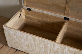 The Aranya Cotton & Jute Upholstered Storage Bench by nkuku features a hinged lid, two spacious interior sections, and textured beige fabric. This stylish bench sits on a light wooden floor beside a light-colored wall.