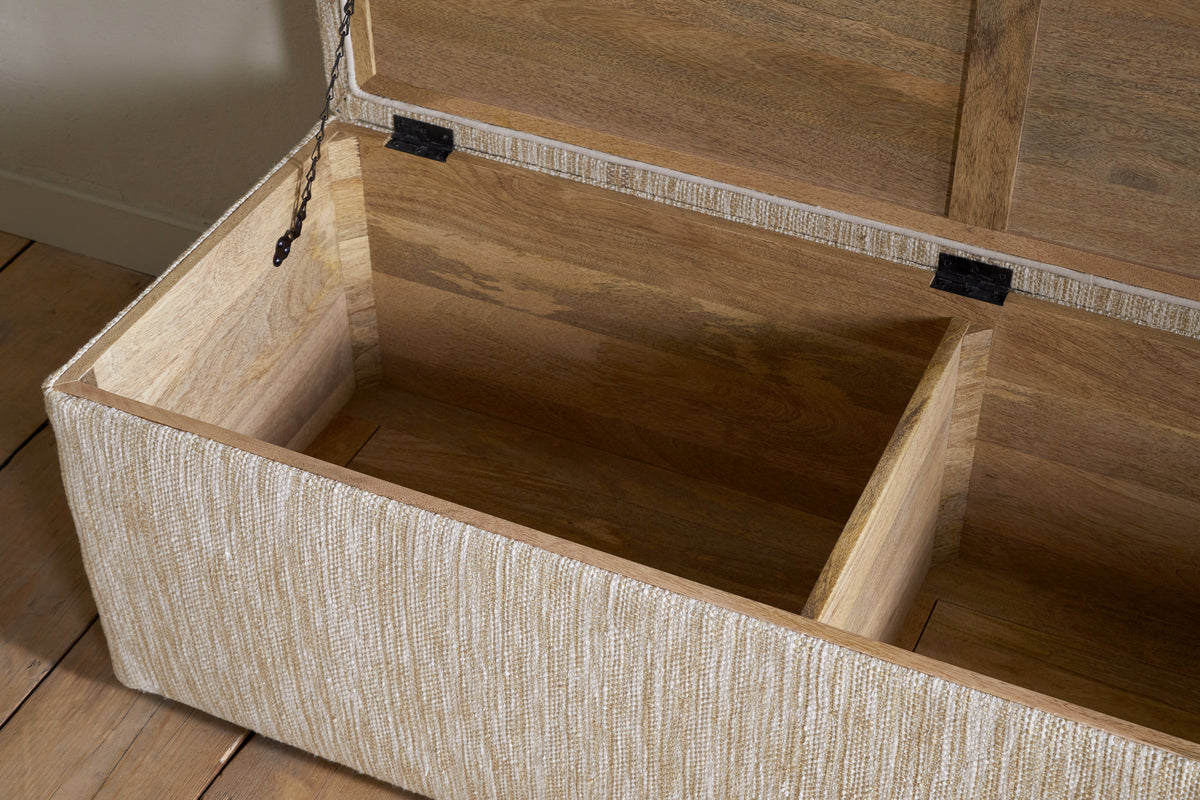 The Aranya Cotton & Jute Upholstered Storage Bench by nkuku features a hinged lid, two spacious interior sections, and textured beige fabric. This stylish bench sits on a light wooden floor beside a light-colored wall.