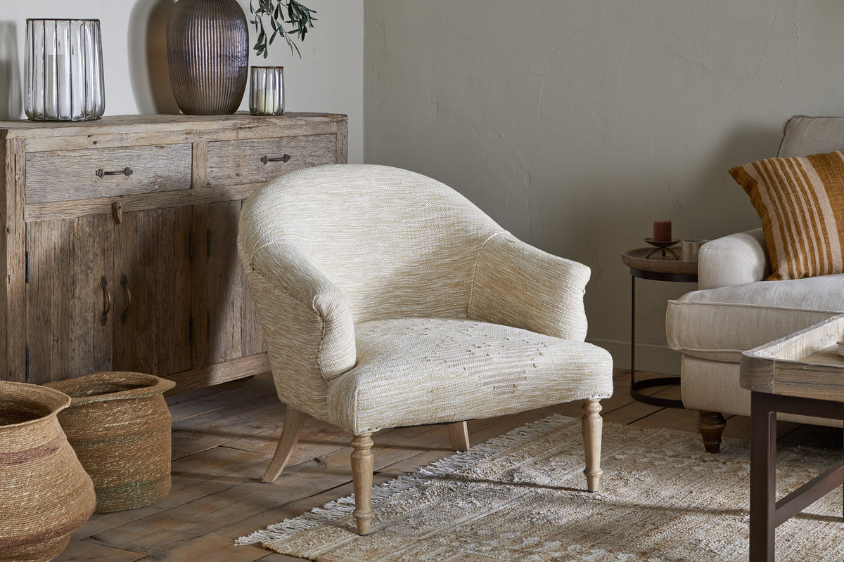 A cozy living room featuring the nkuku Aranya Cotton & Jute Occasional Chair in Natural, wooden legs, a neutral rug, rustic wooden sideboard, woven baskets, and a beige sofa with striped pillows. Earthy decor and soft lighting add warmth.