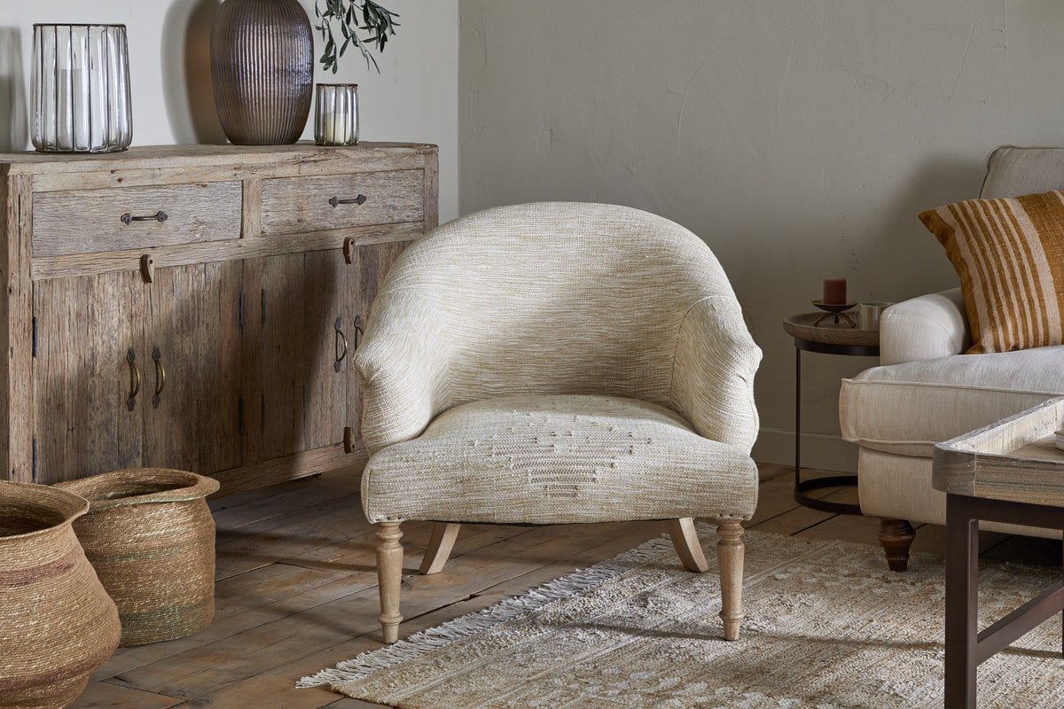 A cozy, neutral living room features nkuku's Aranya Cotton & Jute Occasional Chair in Natural with wooden legs and a supportive seat, set on a fringed rug. A rustic sideboard with glass vases, woven baskets, a beige sofa, and a candle complete the decor.