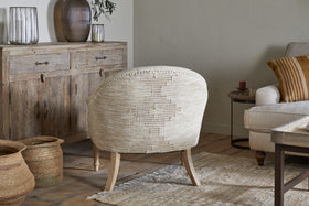 A warm, inviting living room with the nkuku Aranya Cotton & Jute Occasional Chair in natural beige, a rustic wooden sideboard with ribbed glass vases, woven baskets on wood flooring, a cream sofa with striped cushions, and a textured cream rug.