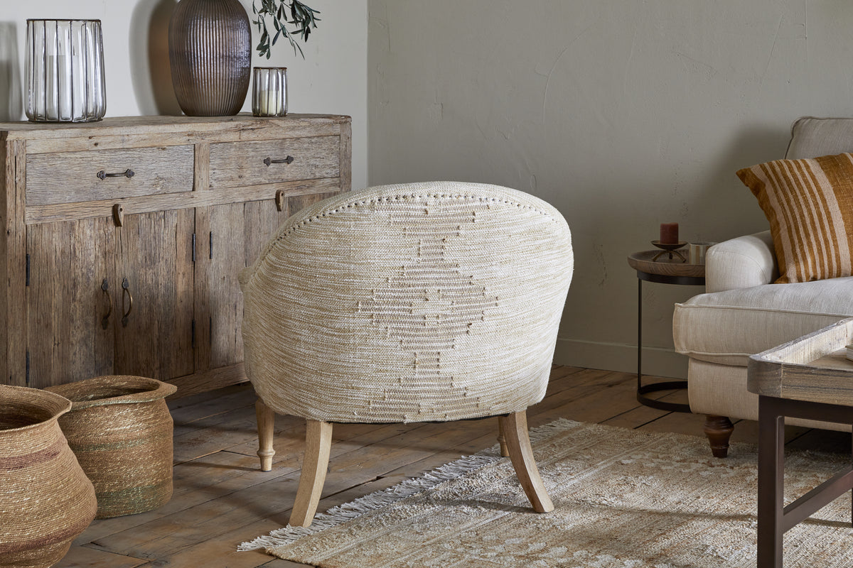 A warm, inviting living room with the nkuku Aranya Cotton & Jute Occasional Chair in natural beige, a rustic wooden sideboard with ribbed glass vases, woven baskets on wood flooring, a cream sofa with striped cushions, and a textured cream rug.