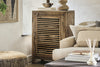 The nkuku Ibo Reclaimed Wood Slatted Low Cabinet - Natural stands by a beige sofa with neutral pillows, holding stacked plates inside and vases on top. A woven ottoman, books, and diffuser sit on a dark coffee table in the bright, cozy room.