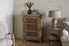 The Ibo Reclaimed Wood Slatted Low Cabinet by nkuku sits on a wooden floor with small plants in dark vases on top. A round side table holds a woven lamp and basket, framed by two beige armchairs for a cozy Indian craft-inspired scene.