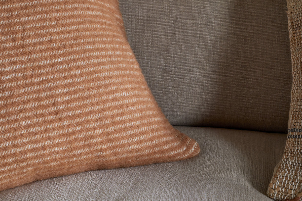 A close-up of the Ilaa Wool Cushion Cover - Sand by nkuku rests on a beige couch, paired with another tan woven pillow to the right. The cozy, neutral-toned setting highlights the cushion's soft brown and white stripes.