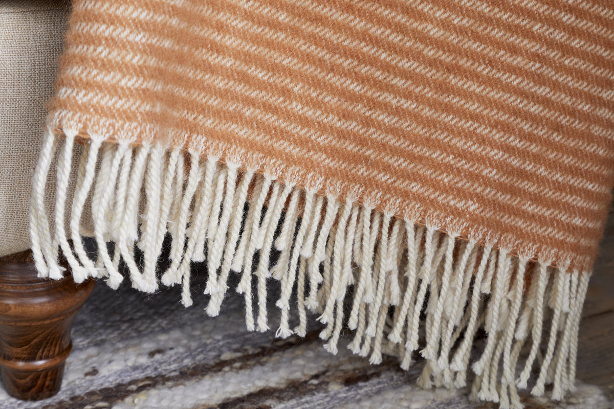 A close-up of the Ilaa Wool Throw - Sand by nkuku, draped over a beige upholstered seat with dark wooden leg; its woven fringes rest atop a textured light rug, highlighting the throw's soft orange-and-cream stripes.