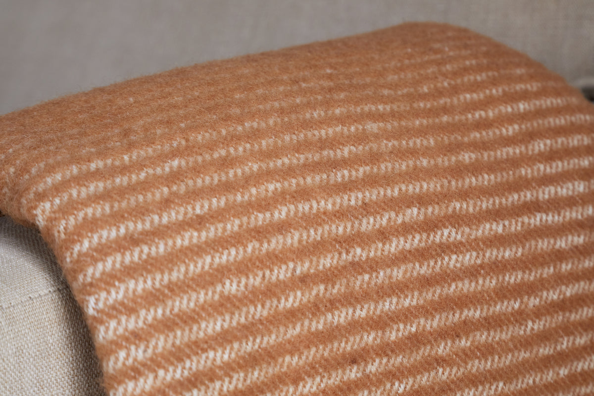 A close-up of the nkuku Ilaa Wool Throw - Sand, draped over a light beige couch. This warm caramel blanket features thin, evenly spaced white stripes across its soft, textured surface.