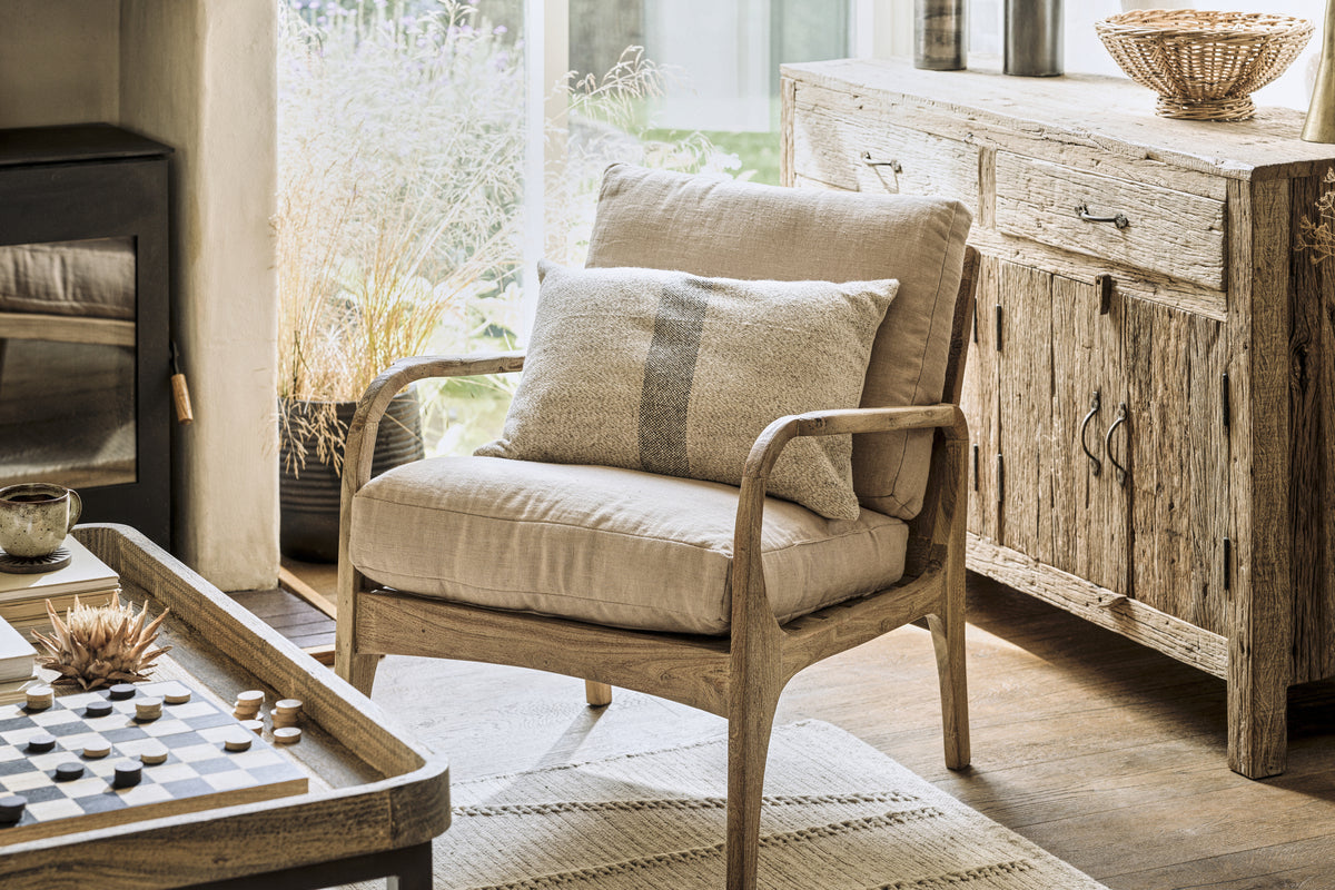 A cozy living room showcases the nkuku Jaya Acacia Wood & Linen Occasional Chair in natural, paired with a rustic sideboard and a sunlit coffee table set with a checkers board and decor.