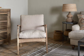 A cozy living room with nkuku’s Jaya Acacia Wood & Linen Occasional Chair - Natural, a striped rug on wooden floors, a wooden cabinet, beige sofa with linen cushions, two woven baskets, round side table, and lamp with woven shade against a light wall.