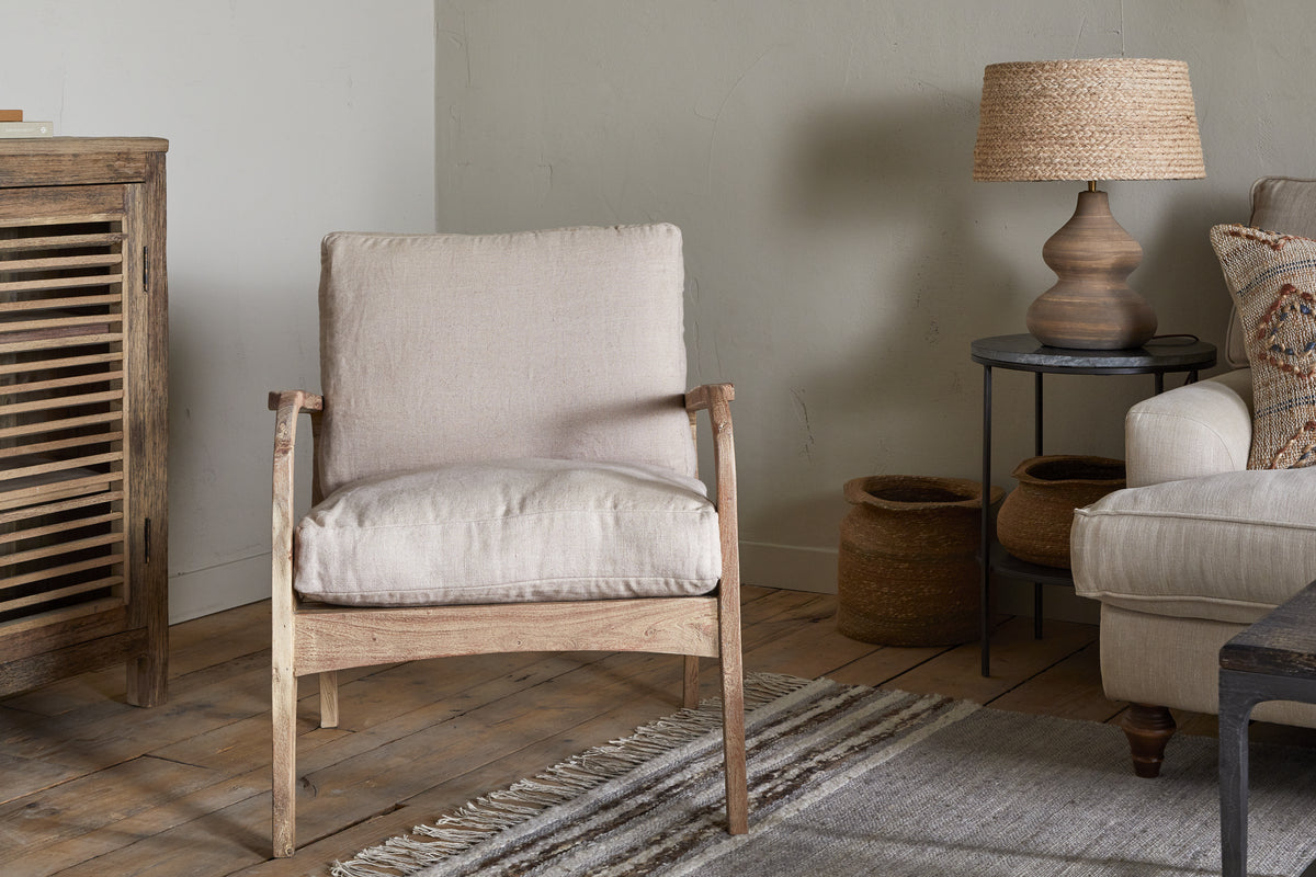 A cozy living room with nkuku’s Jaya Acacia Wood & Linen Occasional Chair - Natural, a striped rug on wooden floors, a wooden cabinet, beige sofa with linen cushions, two woven baskets, round side table, and lamp with woven shade against a light wall.