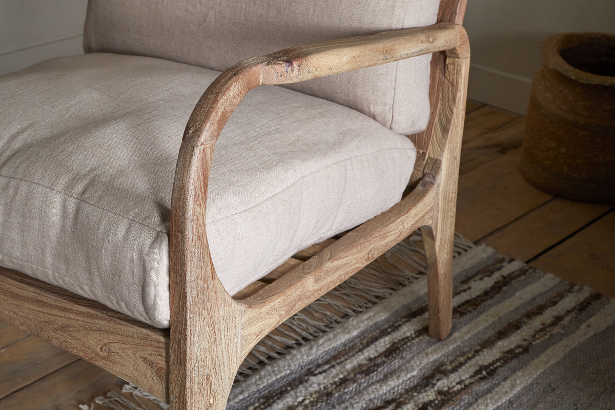 A close-up of the nkuku Jaya Acacia Wood & Linen Occasional Chair in natural shows its curved armrests and light beige linen cushions, set on a striped woven rug with a basket in the background atop a wooden floor.