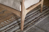 Close-up of the nkuku Jaya Acacia Wood & Linen Occasional Chair in Natural, featuring acacia wood legs and armrest with a beige linen cushion, set on an earth-toned striped woven rug atop a natural wood plank floor.