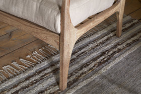 Close-up of the nkuku Jaya Acacia Wood & Linen Occasional Chair in Natural, featuring acacia wood legs and armrest with a beige linen cushion, set on an earth-toned striped woven rug atop a natural wood plank floor.