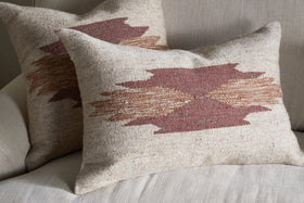Two square throw pillows with nkuku's Kabir Wool Cushion Cover - Rust rest on a light sofa. Each features a geometric pattern in earthy red and brown tones, with overlapping zigzags forming an abstract central design.