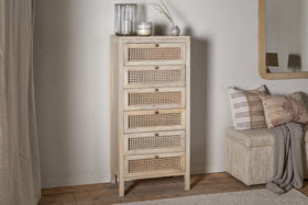 The nkuku Kanz Mango Wood Tall Boy Chest of Drawers - Natural, with six rattan cane drawers and brass knobs, stands against a white wall beside glass vases, a candle, a beige bench with textured pillows, and a mirror on wooden flooring.