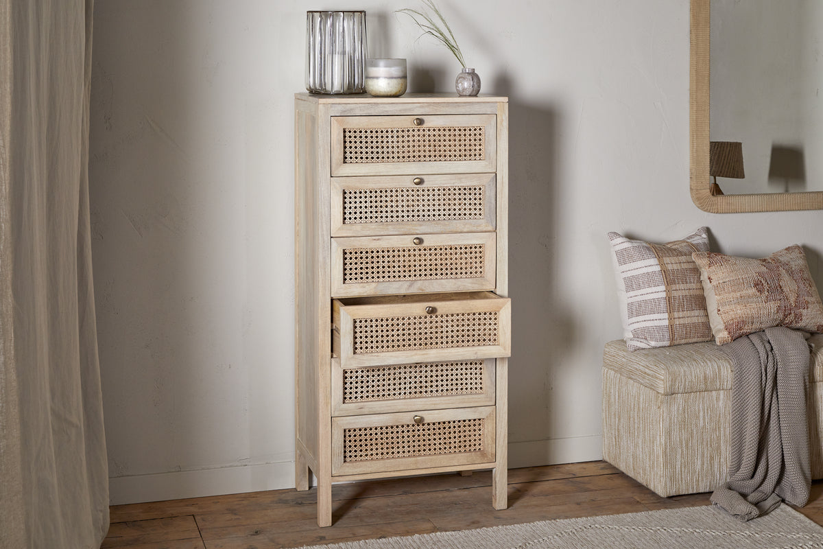 The nkuku Kanz Mango Wood Tall Boy Chest of Drawers - Natural stands on wooden flooring beside a cushioned bench and a textured wall mirror, with glass vases and a candle on top for added charm. Three of its five cane drawers are partially open.