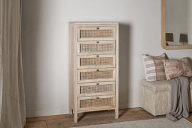 The nkuku Kanz Mango Wood Tall Boy Chest of Drawers - Natural has six cane drawers with round metal knobs and short legs, standing beside a cushioned bench with patterned beige pillows, a draped throw, and a mirror above.