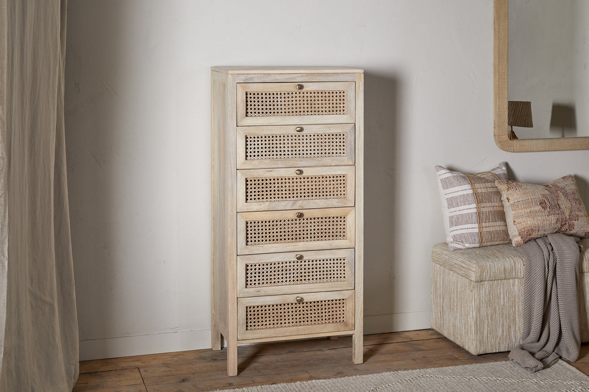 The nkuku Kanz Mango Wood Tall Boy Chest of Drawers - Natural has six cane drawers with round metal knobs and short legs, standing beside a cushioned bench with patterned beige pillows, a draped throw, and a mirror above.