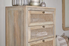 The nkuku Kanz Mango Wood Tall Boy Chest of Drawers - Natural features three cane drawers with round brass knobs. On top are two glass vases, a white candle, and a small vase with reeds. Nearby are a plaid pillow and a partial wall mirror.