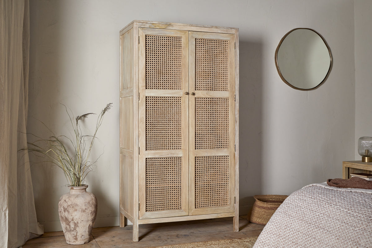 The Kanz Mango Wood Wardrobe by nkuku, featuring natural wood and woven rattan doors, stands against a white wall. A clay vase with dried grasses is on the left; to the right, a round mirror hangs above a wooden nightstand near the bed.