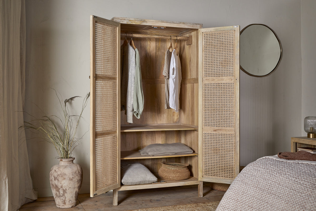 The Kanz Mango Wood Wardrobe - Natural by nkuku stands open, showcasing hanging clothes, folded blankets, and a basket inside. A large vase with dried grasses and a round mirror complete the cozy, softly lit space.