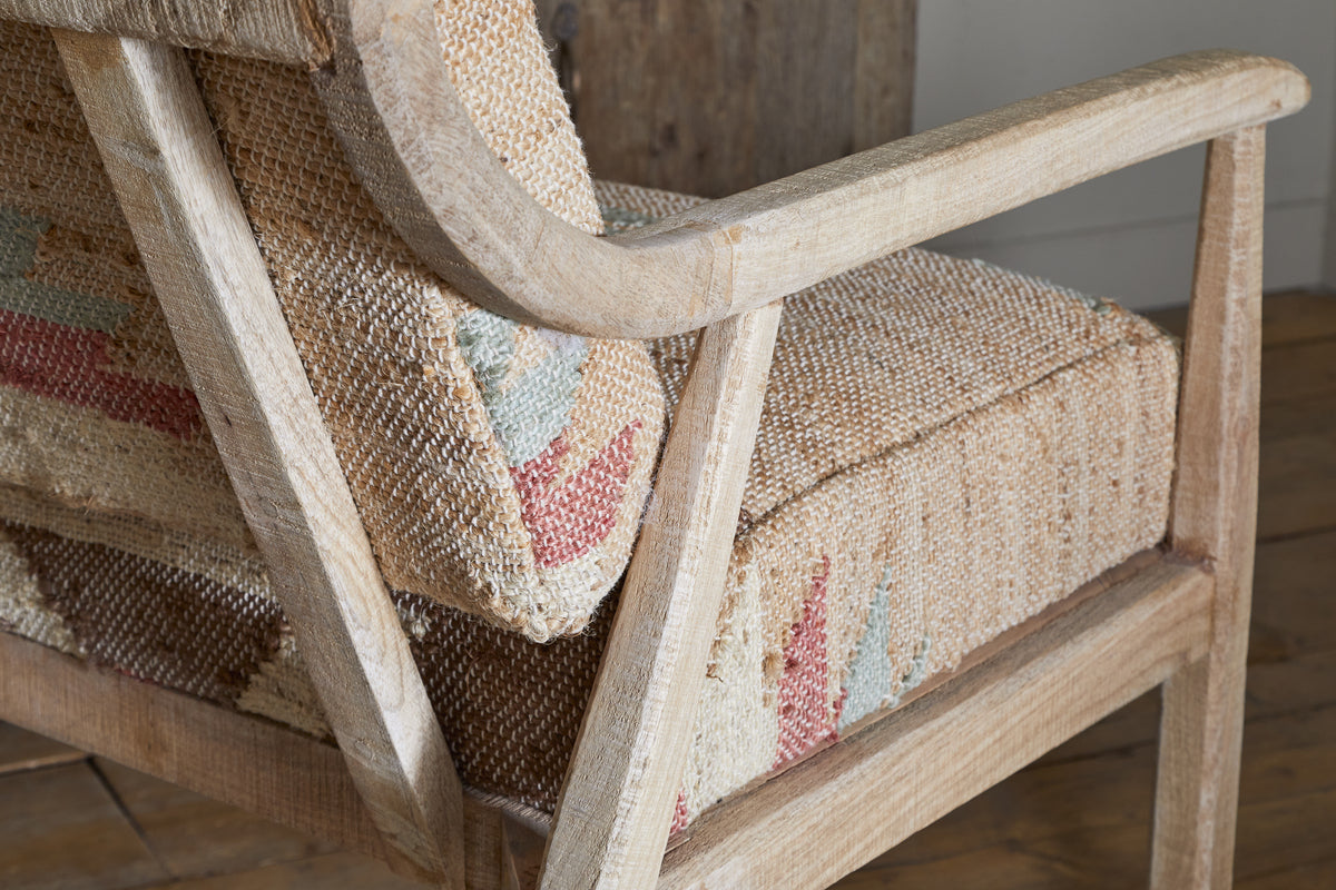 The Kavya Kilim Occasional Armchair by nkuku features a natural, weathered mango wood finish and textured cushions with red, green, and beige patterns. Perfect for rustic spaces, this armchair adds warmth beside any wooden floor or wall.