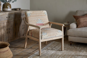 The nkuku Kavya Kilim Occasional Armchair - Natural, with beige and pastel geometric upholstery, sits on a light rug by a cream sofa. Behind the mango wood chair is a wooden sideboard with decor and a woven basket on the floor.