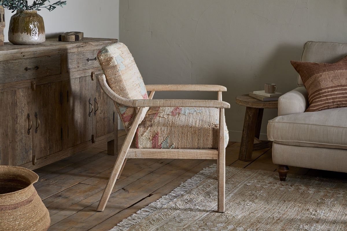 The nkuku Kavya Kilim Occasional Armchair - Natural, featuring a patterned cushion, sits on a patterned rug beside a beige sofa with brown pillows, a rustic wooden side table, and a large wooden sideboard in a cozy, sunlit living room.