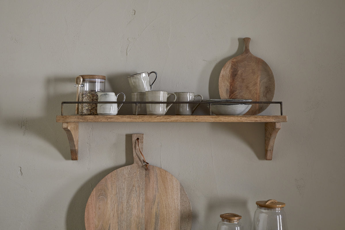 The nkuku Meera Mango Wood & Iron Gallery Shelf - Natural displays ceramic mugs, bowls, a glass jar with wooden lid, and a mango wood cutting board. Below, a round wooden board hangs on the wall in this minimal, neutral-toned scene.